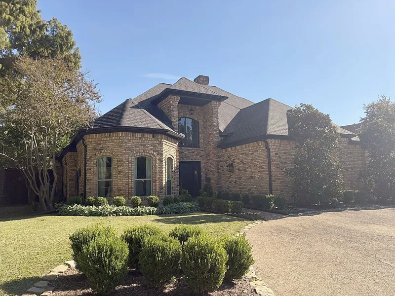A brick two-story home with a turret and a dark shingle roof behind a row of shrubs