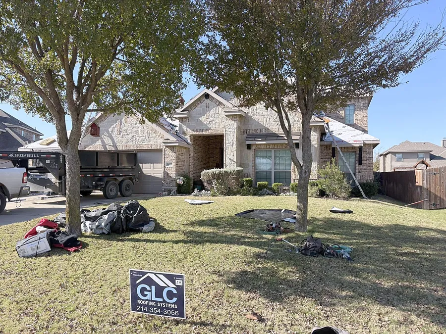 A roof replacement under way on a stone and brick two-story home, with a dump trailer in the drive and the GLC sign on the lawn
