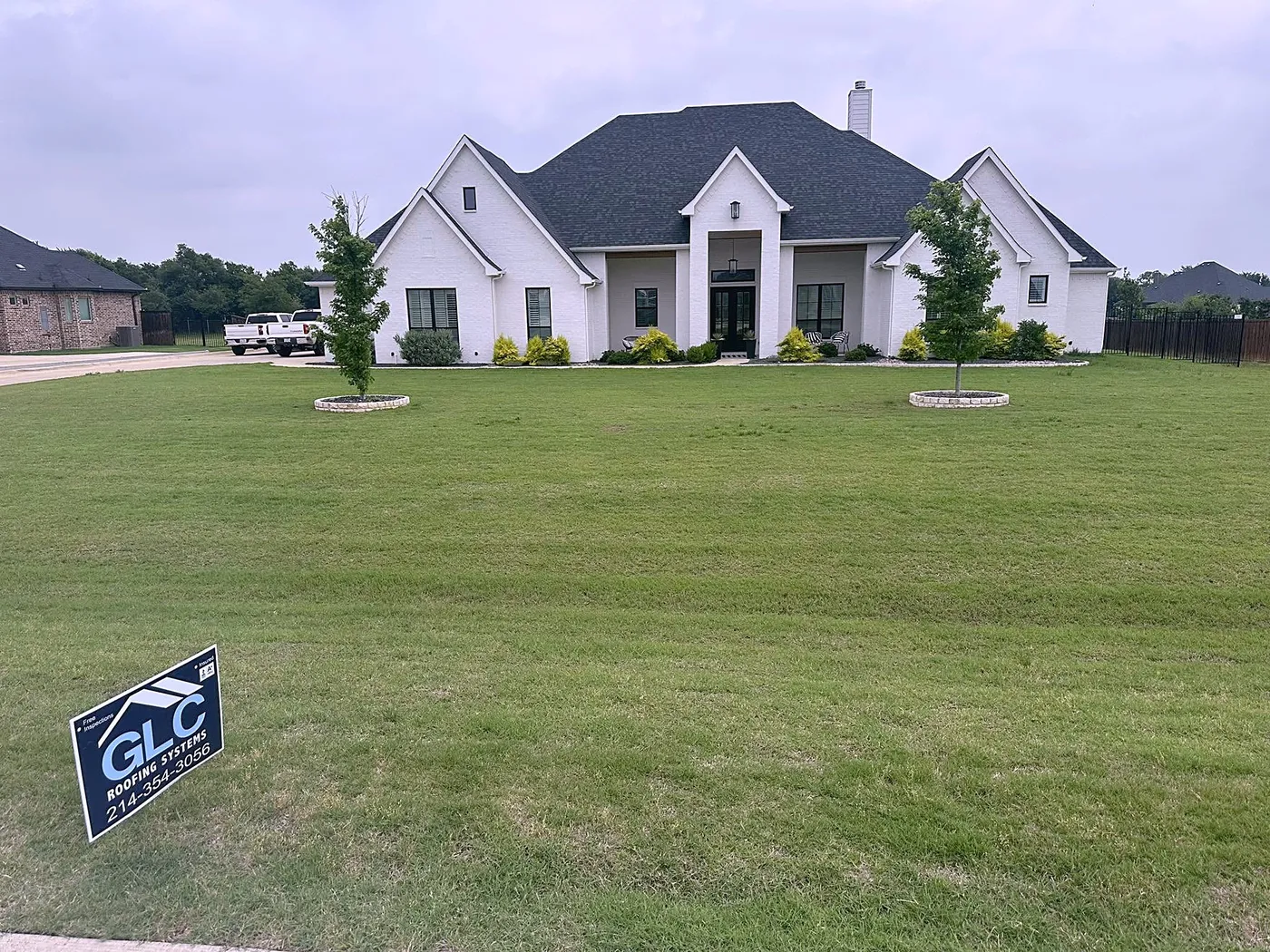A white two-story home with a new dark shingle roof and the GLC Roofing Systems sign on the lawn